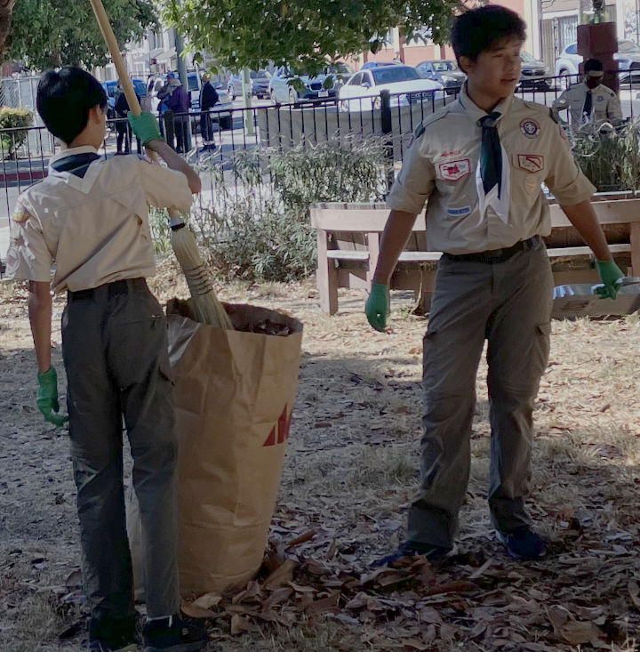 A scout compresses leaves in a bag and another scout picks up trash in the background