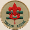 Troop guide badge: two horizontal green lines with a red scouting symbol on top and the words troop guide curved near the edge of the bottom semicircle