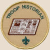 Troop historian: an open book with a small trefoil below it and the words troop historian curved near the edge of the top semicircle