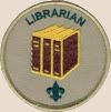 Librarian badge: the word library curved near the edge of the top, three golden books with brown spines and covers, and a small trefoil near the bottom